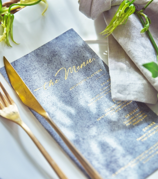 ゴールドのカトラリーとグレーのメニュー表が並ぶ洗練されたテーブルコーディネート